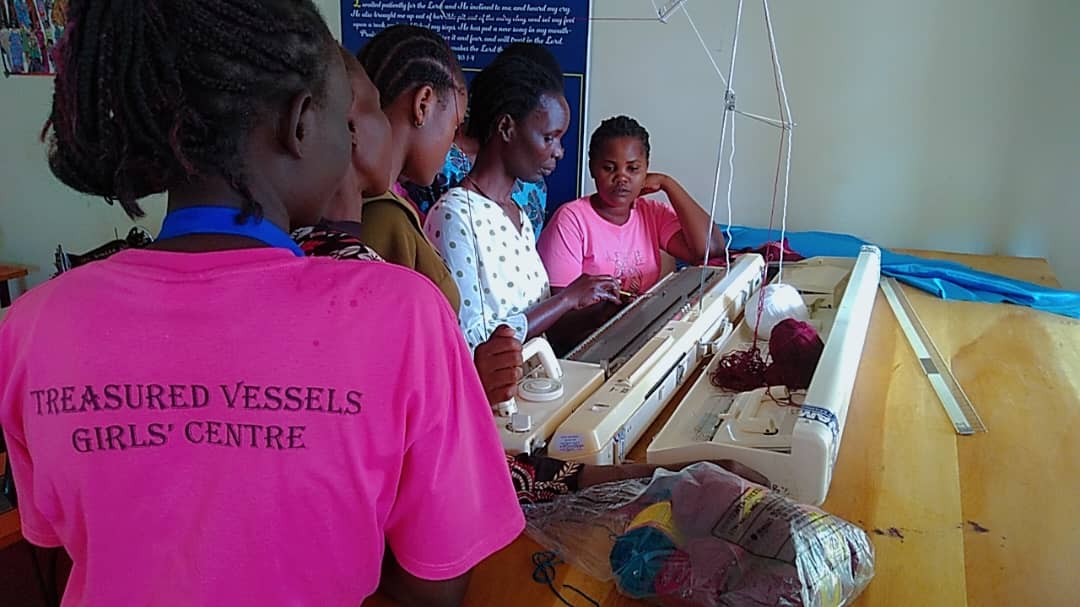 A trainee in a Treasured Vessels Girls' Centre shirt working at a knitting machine in Jinja, Uganda